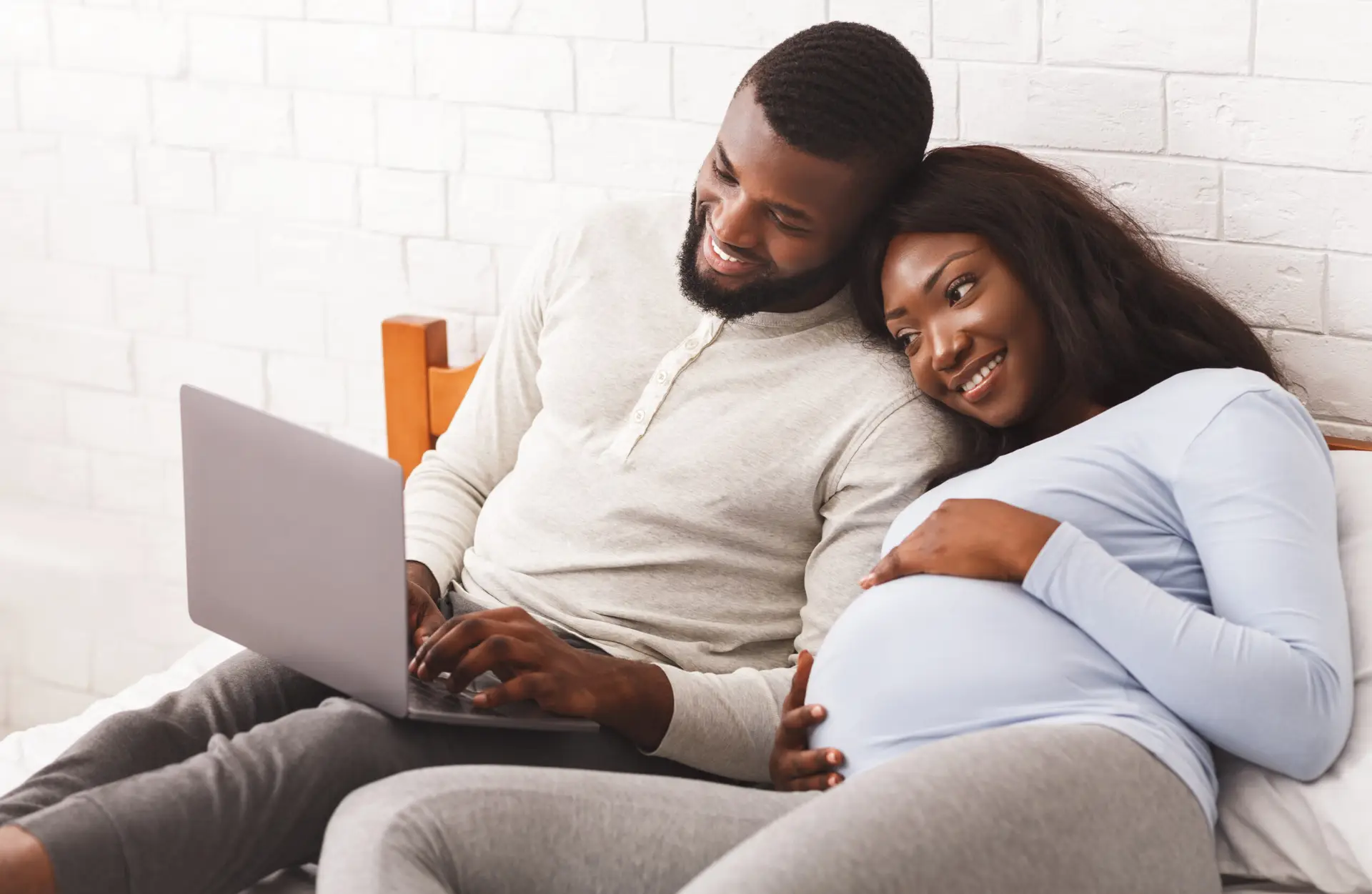 Lovely pregnant afro couple using laptop while resting at home, laying on bed, woman leaning on man shoulders, copy space